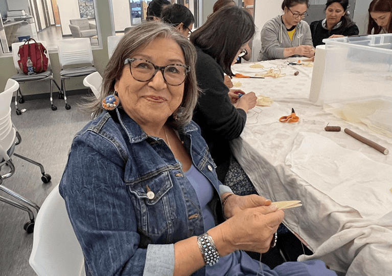 Evelyn Good Striker sitting in a workshop meeting area, smiling at the camera working on a crafting project